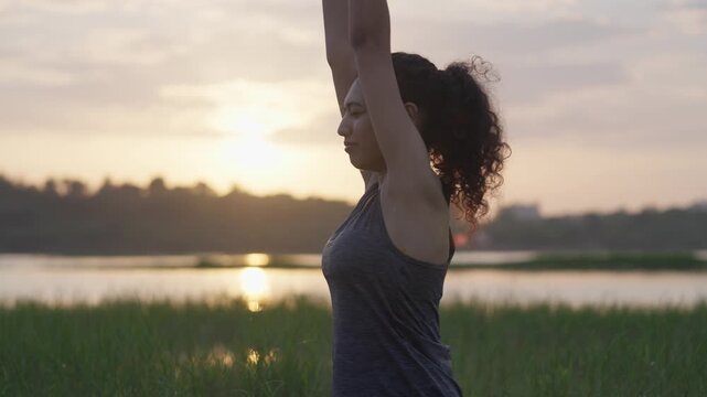 A South Asian woman practices yoga near a lake at sunrise, standing in a peaceful prayer position