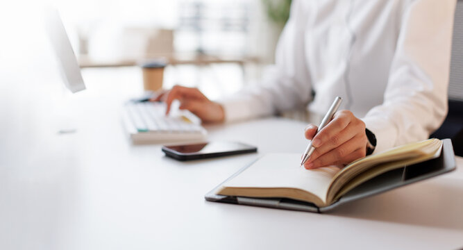 Hands multitasking at a bright desk, writing in a notebook while typing on a laptop keyboard, conveying focused planning, productivity and organized modern work life
