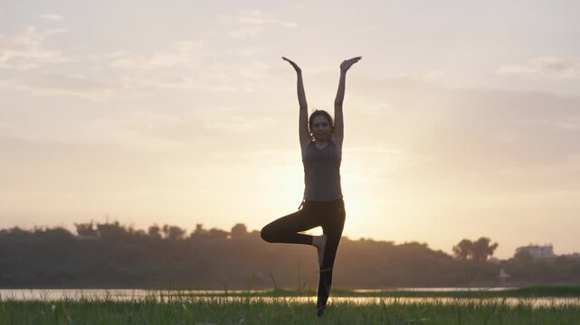 A wide-angle shot of a South Asian woman performing a balanced tree pose on a grassy lakeside at sunrise, silhouetted against the golden sky