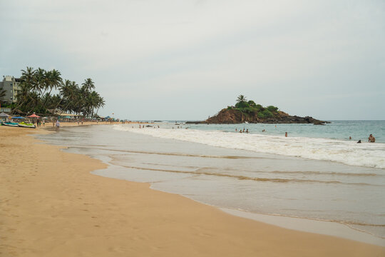 View of Mirissa Beach with people swimming near Parrot Rock and palm trees lining the shore in Mirissa, Southern Province, Sri Lanka.
