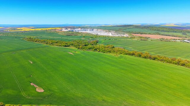 Panorama of green agricultural landscape with distant stone quarry. Wide aerial view of vast vibrant green fields under clear blue sky with industrial mining quarry on horizon.