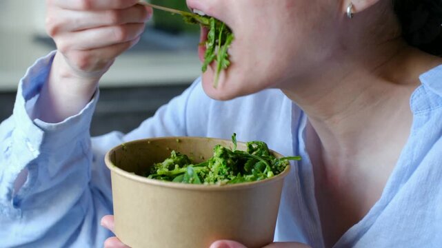 Close up of a young woman enjoying a healthy green salad from a craft paper takeaway bowl, using a wooden fork for a conscious and sustainable lunch break at home or in the office
