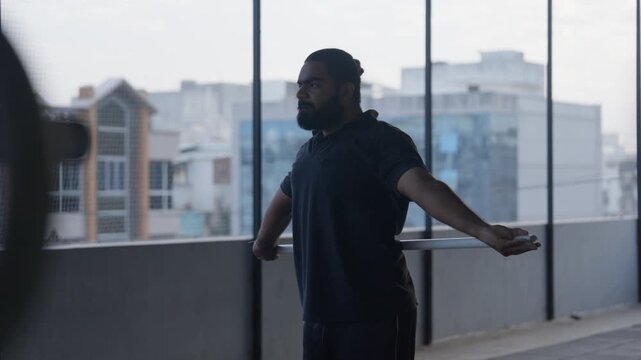 A smooth tracking shot follows a confident Indian bearded man in dark athletic wear as he extends his arms wide in a dramatic windowed modern gym overlooking a city skyline. Cinematic right-to-left ca