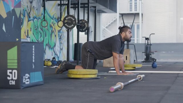 A powerful Indian bearded athlete performs dynamic bear crawl warm up exercise on the floor of a vibrant CrossFit gym with a striking colorful graffiti mural wall. Side profile static shot captures fu