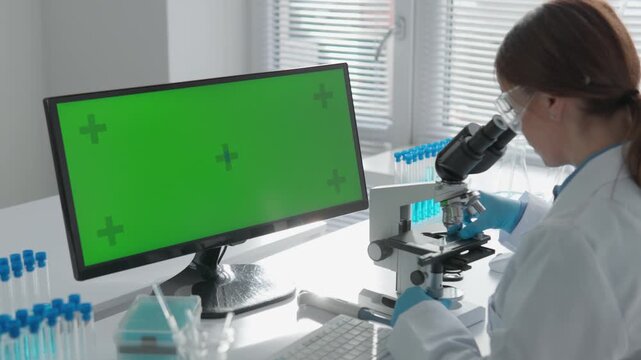 A researcher conducts scientific analysis using a microscope while a green screen computer monitor displays in the background. Laboratory glassware and natural light enhance the innovative workspace