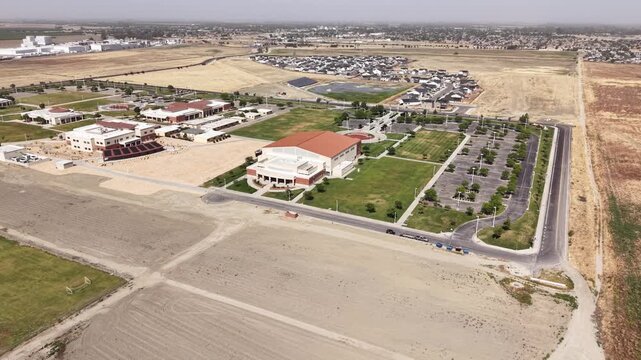 High-angle panning view of the Lemoore College campus, showing modern university buildings, an expansive parking lot, and surrounding rural landscape in Central California during a sunny day