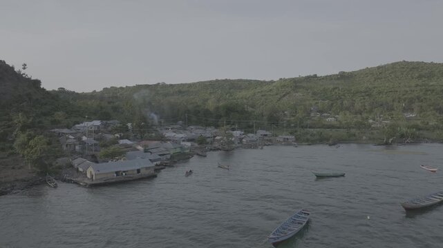 Low angle drone shot of a densely populated coastal fishing village built along the shoreline, with clustered corrugated iron rooftops, wooden fishing boats docked in a small harbor inlet sunsetting