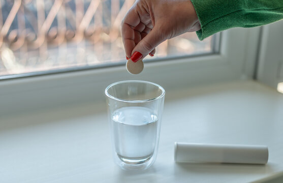 Woman dropping dissolvable tablet into glass of water near window, concept of hydration, vitamins, healthcare and morning wellness routine