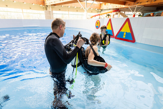 Scuba diving instructor helping a teenage girl put on her dive gear in the shallow water of an indoor pool