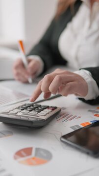 Closeup of female hands calculating data and reviewing business analytics on printed charts. Office workspace with calculator, smartphone, and financial documents for budget analysis and planning