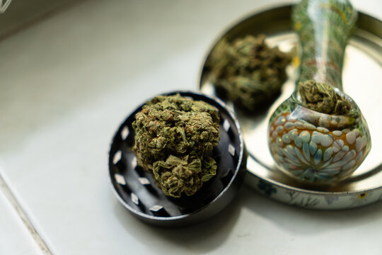 Overhead view of a large dry weed bud, an open metal grinder, and a glass smoking pipe inside a stash tin.