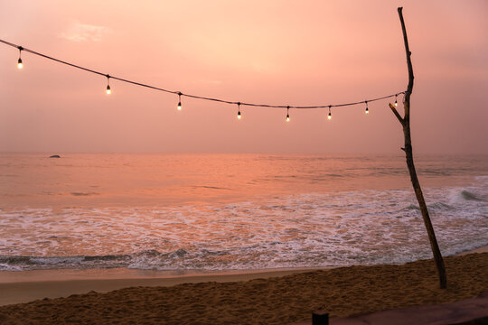 View of a beach at sunset with a string of glowing decorative lights hanging from a wooden pole over the sandy shore, Galle, Southern Province, Sri Lanka.