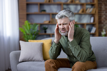 Fototapeta premium Mature man sitting on a sofa at home, experiencing intense headache or migraine pain, holding hands to temples with a pained expression, symbolizing stress, health issues, and discomfort