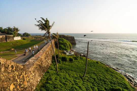 View of the historic Galle Fort ramparts with people walking along the stone walls, leaning palm trees, and the Indian Ocean coastline in Galle, Southern Province, Sri Lanka.
