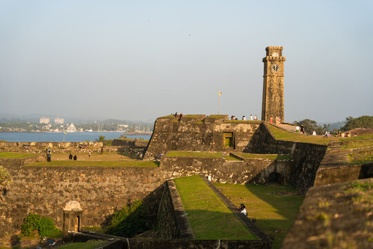 View of the historic Galle Fort Clock Tower and stone ramparts with people walking along the fortifications under a clear sky in Galle, Southern Province, Sri Lanka.