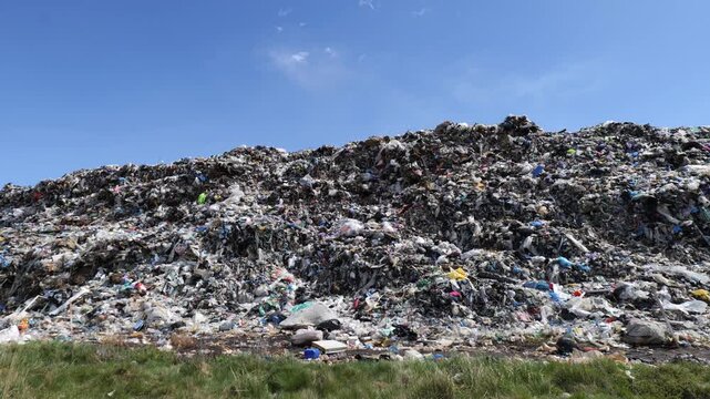 The garbage dump fills with various items and trash under a clear sky as people walk nearby. Dumping continues in this area