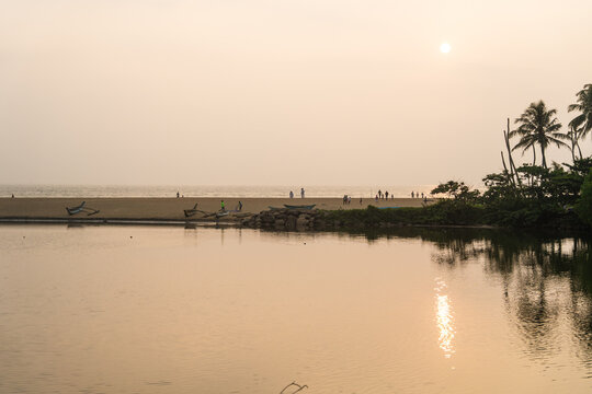 View of traditional outrigger boats and people on a sandy beach by calm water at sunset Galle, Southern Province, Sri Lanka.