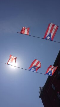 A string of Puerto Rican flags waving in the wind against a clear blue sky with sun rays.
