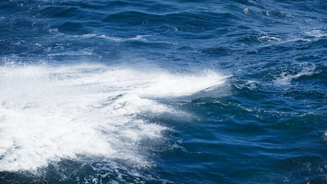 White foam spreads across deep blue water. Ocean surface shows waves, spray, turbulence and marine texture. Ocean creates dynamic seascape with movement and aquatic energy