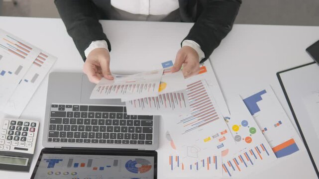 Top view of a person working with colorful business reports, pie charts, and bar graphs on a white desk. Modern workplace setup highlights financial analysis, productivity, and digital workflow
