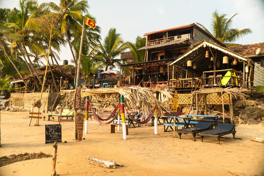 View of beachfront bars and restaurants with thatched roofs, hammocks, and palm trees on a sandy shore in Galle, Southern Province, Sri Lanka.