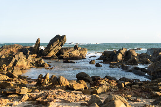 View of large dark rock formations and boulders scattered along the sandy shoreline with ocean waves crashing against the coast under a clear sky in Galle, Southern Province, Sri Lanka.