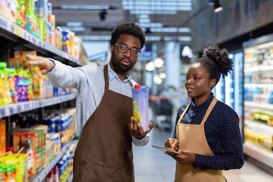 Experienced male grocery store manager teaching a female colleague about product placement and inventory control, pointing to items on shelves while she takes notes on a tablet