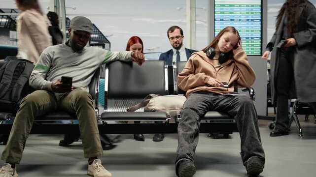 Candid airport terminal scene with a traveler crossing the frame in motion blur while a seated passenger waits near a backpack and departure screens in the background.