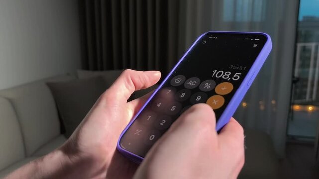 Hands use a smartphone calculator in close up. The mobile device enters numbers on a dark screen in a living room. App finance, budgeting, and technology mockup..
