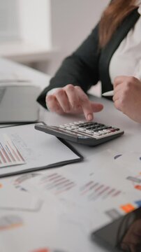 Closeup of female hands calculating numbers on a calculator with financial charts and paperwork on the table. Office environment focused on accounting, finance analysis, and business strategy planning
