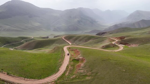 A lone car winds along a serpentine dirt road through Kazakhstan&rsquo;s sweeping green highlands, framed by layered mountain ridges fading into the haze. This cinematic aerial establishes a powerful sense