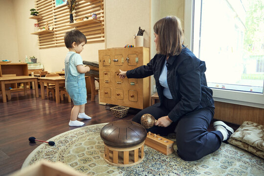 Toddler exploring wooden activity board while educator guides fine motor learning. Concept of childcare support, sensory play, preschool development, speech practice, and patient guidance.