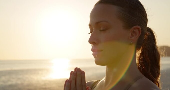 Portrait of dedicated young woman shows namaste mudra with hands on sunrise beach closeup. Relaxed female practices yoga and meditation at tropical resort