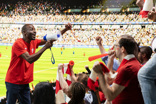 Black capo leads synchronized stadium chants with megaphone, supporter group celebrating together in the stands. Sports fans with face paint and scarves, cheering and stadium noise.