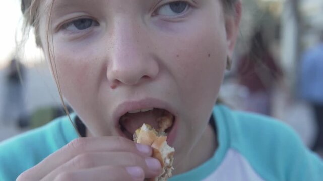 Close-up of a young girl finishing a cheeseburger outdoors on a seaside promenade in the evening. Cropped lifestyle shot showing a child taking the last bite of fast food during a casual walk, with