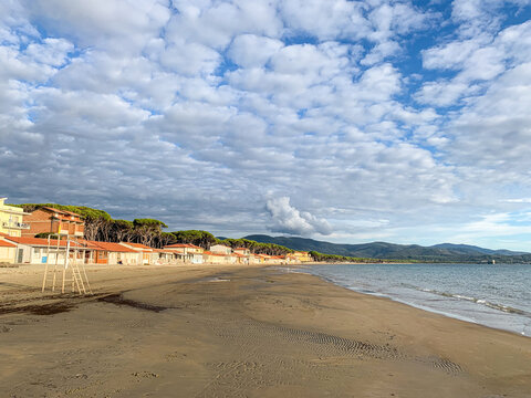 A wide view of the quiet sandy beach in Follonica, Italy, showing coastal houses, green pine trees, calm sea waters, and a dramatic sky filled with scattered clouds.
