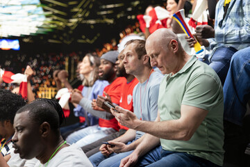 Football supporter placing live betting wagers on mobile gambling app, while watching soccer match in stadium stands. Fan follows sports predictions during intense in-play betting experience. © DC Studio