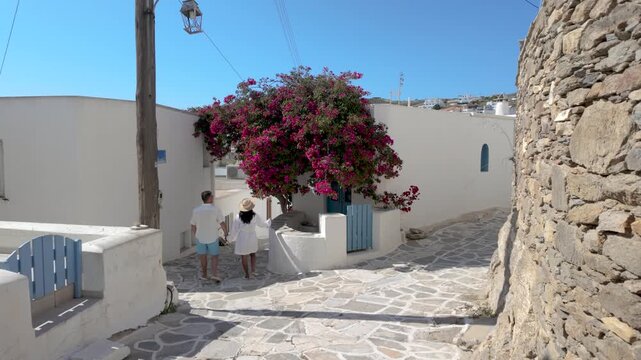 A diverse couple wanders along picturesque pathways in Lefkes, Paros, Greece, surrounded by vibrant bougainvillea. The beautiful architecture and blue accents enhance the enchanting holiday atmosphere