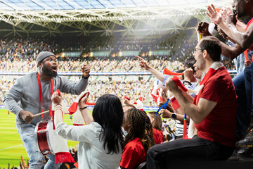 Obraz premium Supporter group leader creating intense energy by playing fan drums and leading chants from fan section during football match. Diverse sports fans with face paint and raised scarves in passion.