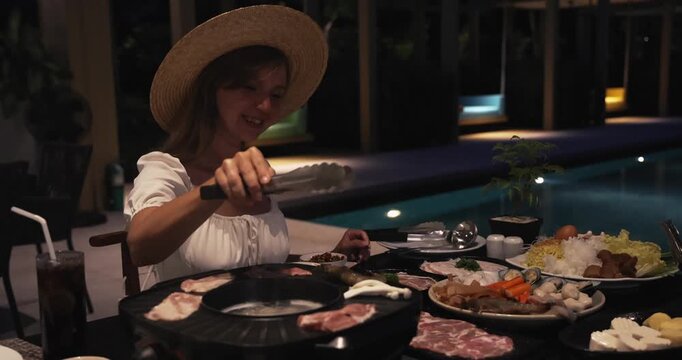 Woman cooking sliced meat on mookata grill at night, poolside resort patio, luxury vacation dining, outdoor lifestyle, Asian BBQ food culture