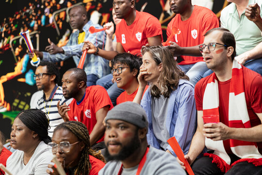 Red team football supporters watching tense live soccer match from stadium stands, mixed emotions, horns and anxious crowd reaction. Sports fans clenching fists speechless after missed goal.