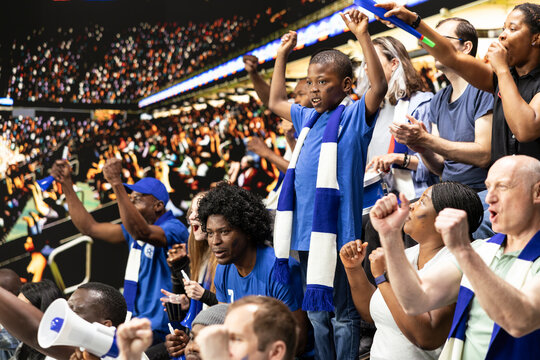 Young soccer fan cheering with blue supporters during football match in packed stadium filled with cheering supporters. Family outing captures fatherhood and role model during soccer experience.