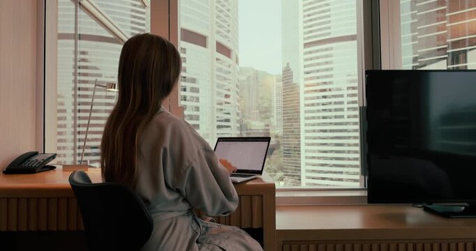 Woman in bathrobe typing on laptop and leaning back in chair to rest in a high-rise hotel room overlooking city skyscrapers during the day. Medium shot.