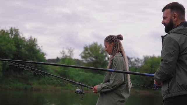 Young couple enjoying a relaxing day fishing together on the riverbank, holding their rods and waiting for a catch while surrounded by serene nature and lush green trees on a cloudy day