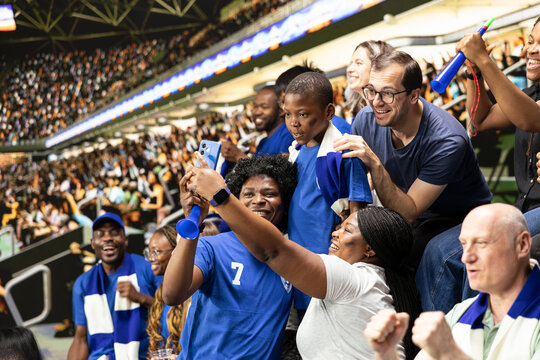 Happy soccer fans taking selfie with child during live match in crowded stadium, family outing during first match with cheering supporters group. Fun sports and crowd excitement.