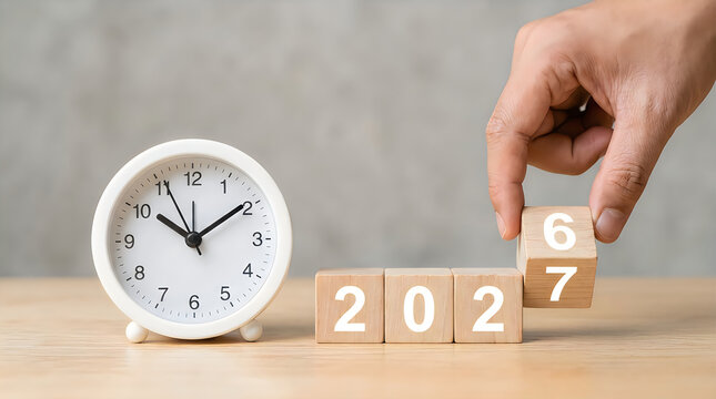 Hand changing wooden blocks from 2026 to 2027 with a clock on the table, concept of time passing and new year countdown