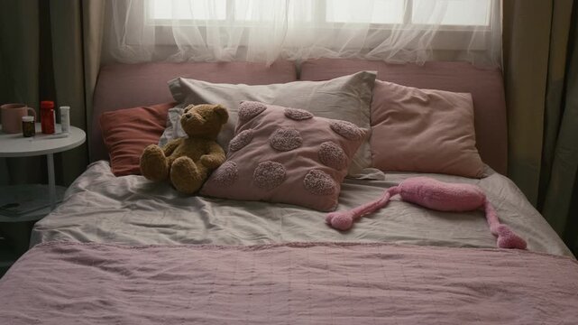 Zoom in shot of cozy bed with pink and grey pillows, plush bear, plush heart-shaped toy near white side table showing pill bottles in dimly lit bedroom