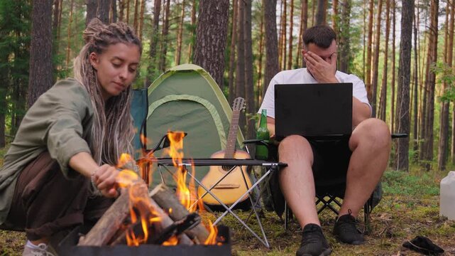 Young couple camping in the forest, where the woman is making a bonfire while her partner, a remote worker, is busy with his laptop before finally closing it to relax with her