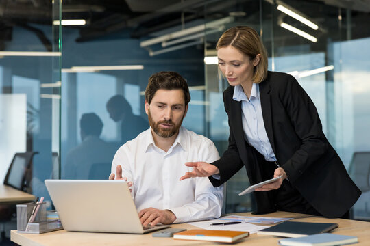 Two business professionals in an office environment discussing a project, with the woman providing guidance or supervision to the man working on a laptop, fostering teamwork and mentorship