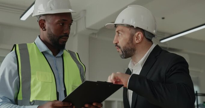 Portrait of Middle Eastern and African American construction managers in helmets discuss site plan. Supervisor speaks with engineer and gives instructions